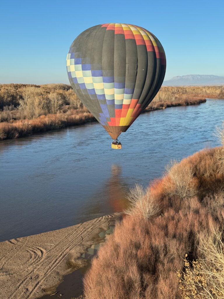 A colorful hot air balloon floats low over a calm river, surrounded by dry, brown brush and trees on a clear day with blue sky and distant mountains—perfect for riding in a hot air balloon and enjoying serene views.