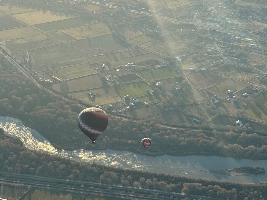 Riding in a hot air balloon, two colorful balloons float above a river bordered by green fields, farmland, and a patchwork of trees, all basking in the sunlight from high above.