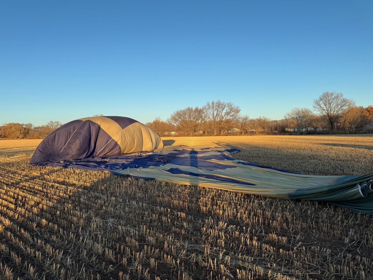 A deflated hot air balloon lies on a harvested field at sunset, its fabric spread across the ground—a quiet ending after riding in a hot air balloon beneath clear blue skies and trees in the distance.