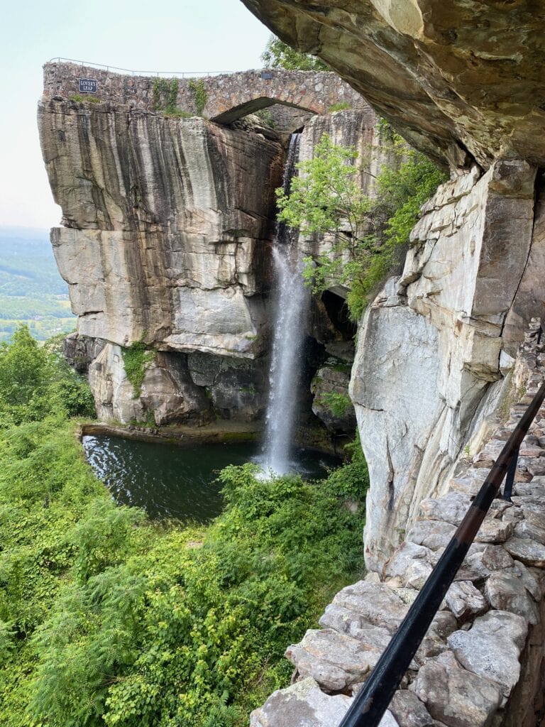 A waterfall cascades from a stone bridge into a pool below, surrounded by lush greenery and rocky cliffs, with a metal railing along a stone pathway in the foreground.