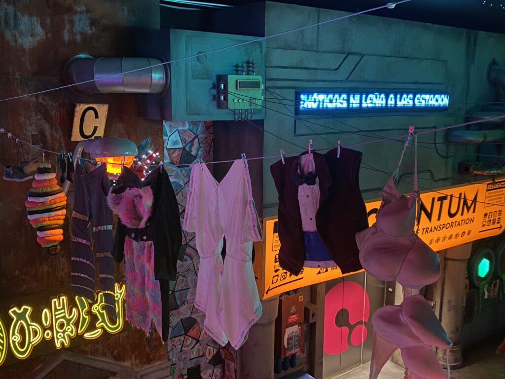 Colorful and eclectic clothing hangs on a clothesline above a futuristic, neon-lit street with bright signs and a glowing blue electronic message board in Spanish. The scene has a sci-fi, cyberpunk aesthetic.