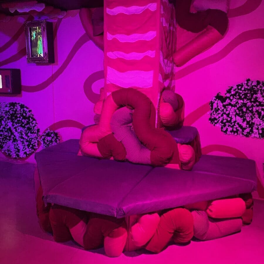 A surreal art installation features a tiered structure covered with plush, intestine-like tubes in shades of pink and red, surrounded by pink walls with wavy patterns and artificial greenery, all illuminated by pink lighting.