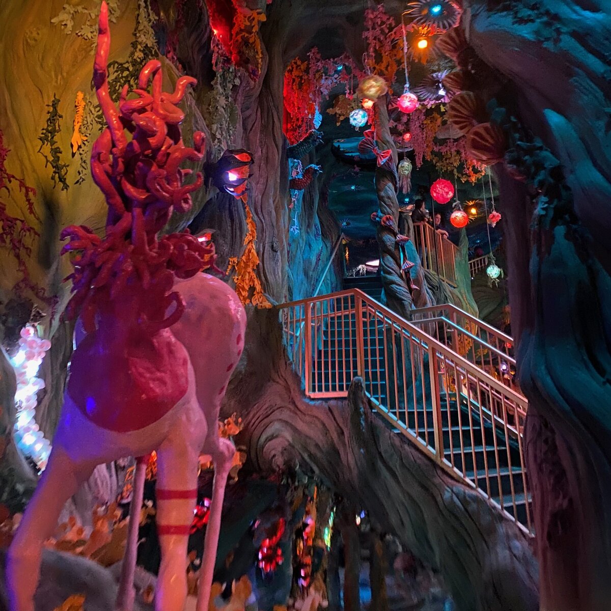 A whimsical, colorful indoor scene features a white unicorn statue, glowing orbs, vibrant lights, twisted staircase, and fantasy-themed decor resembling a magical forest with swirling trees and hanging lanterns.