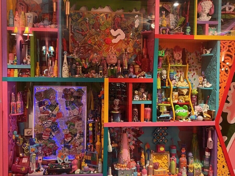 A colorful display case filled with eclectic toys, figurines, and art objects stands against a vibrant wall with a cloud-patterned ceiling and red-and-blue checkered floor. The shelves are packed with playful, whimsical collectibles.