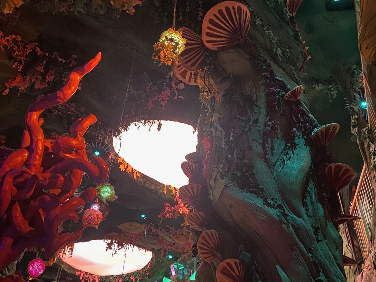 A whimsical, otherworldly indoor scene featuring large artificial trees with pink mushrooms, glowing orbs, and colorful lights creating a fantasy-like atmosphere.