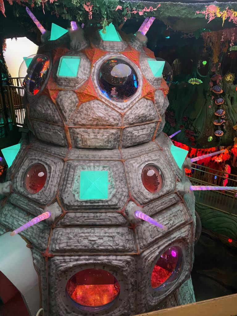 A large, futuristic, gray structure with circular windows and glowing neon green and purple lights, set in an indoor environment with foliage and colorful decorations surrounding it.
