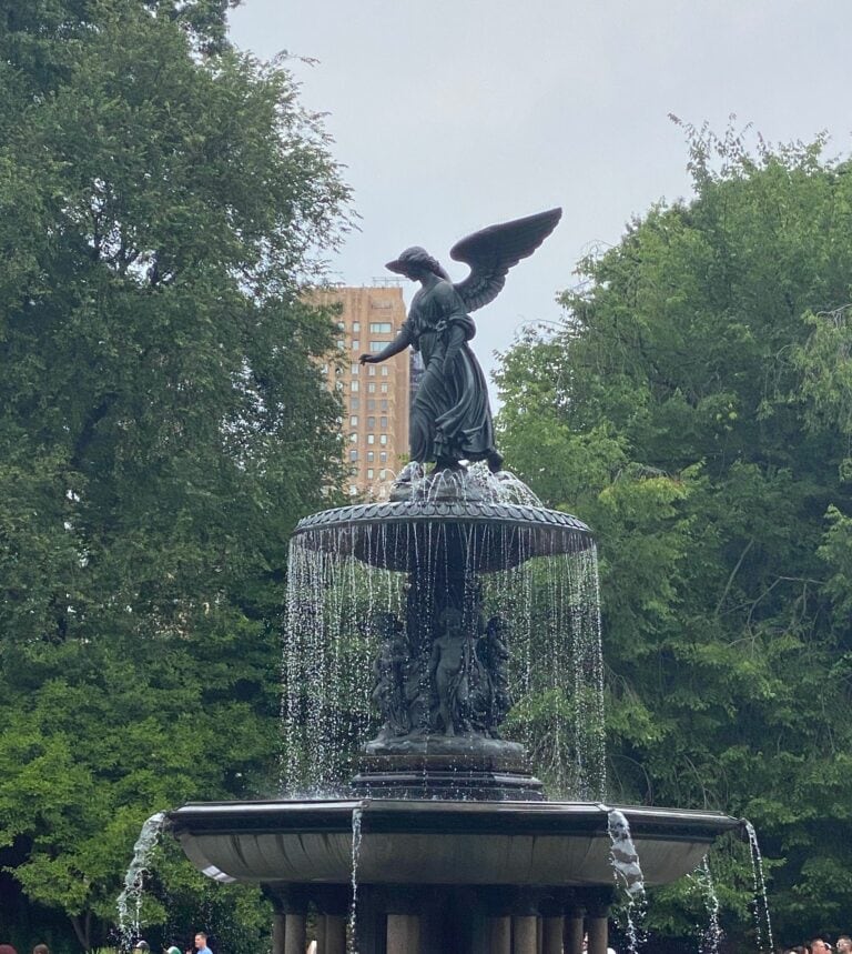 A bronze angel statue stands atop a tiered fountain with water cascading down, surrounded by lush green trees and a tall building visible in the background—a perfect spot for those seeking quiet things to do in NYC.