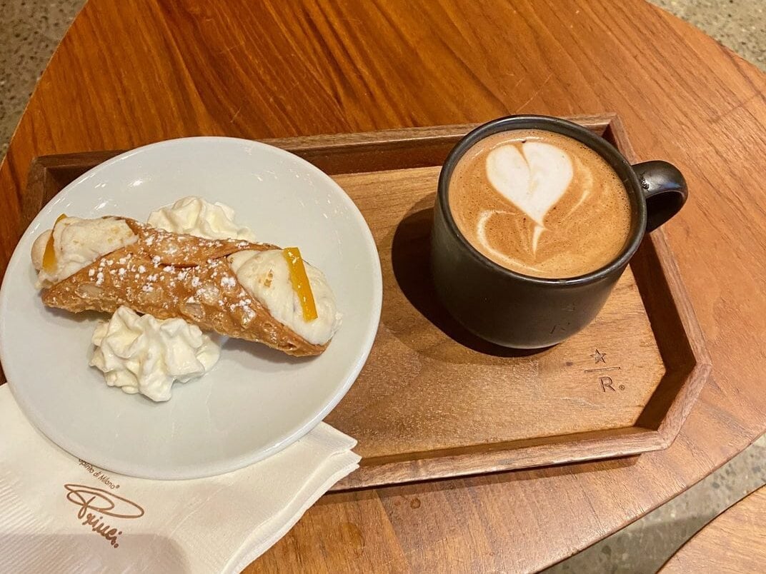 A wooden tray holds a black cup of coffee with heart-shaped latte art and a white plate with a cannoli and whipped cream—perfect for those seeking quiet things to do in NYC, placed on a wooden table with a napkin nearby.