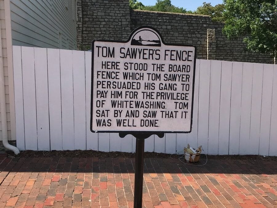 A historical marker in front of a white wooden fence reads: Tom Sawyer’s Fence. Here stood the board fence which Tom Sawyer persuaded his gang to pay him for the privilege of whitewashing. Tom sat by and saw that it was well done.