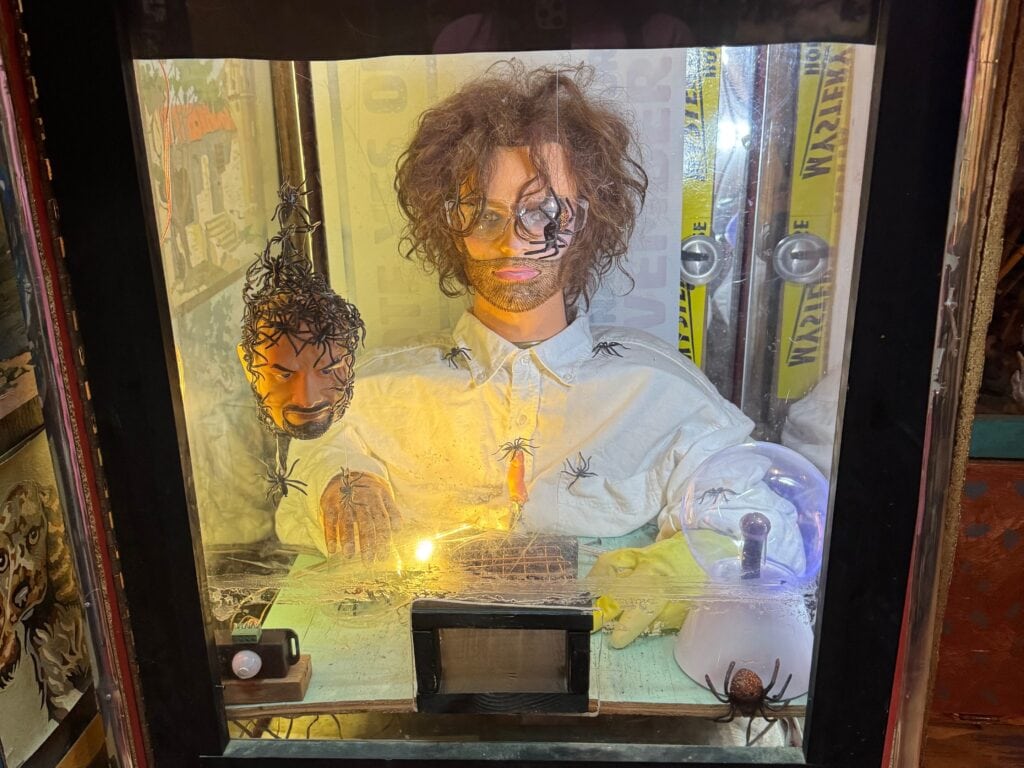 A vintage arcade machine displays a scene with a mannequin scientist and a floating, severed head. The mannequin has disheveled hair, wires, and a spider nearby, with illuminated buttons labeled START and AFTER at the bottom.