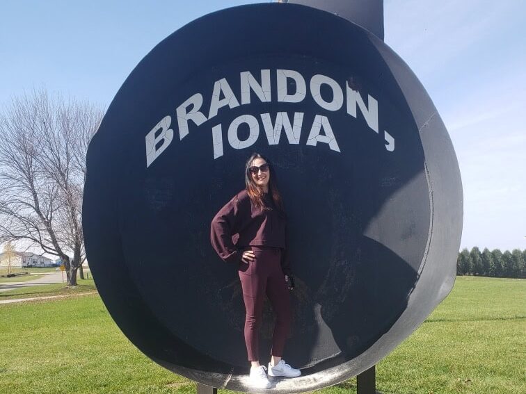 A woman in sunglasses and maroon clothing stands inside a large black metal sculpture with the words BRANDON, IOWA written on it, in a grassy outdoor area on a sunny day.