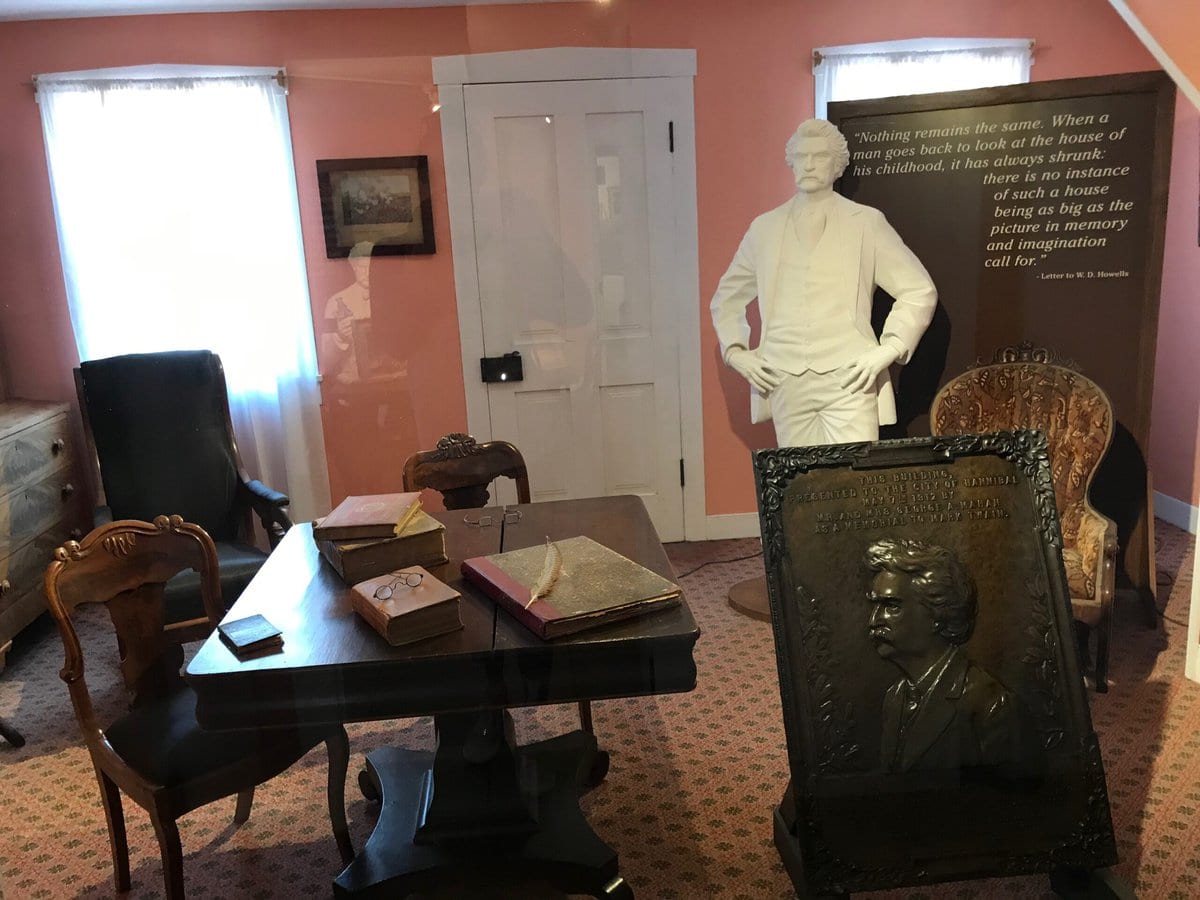 A historical room display features antique wooden furniture, books on a desk, a bust and plaque of Mark Twain, and a white statue of him near a quote on a standing banner, all set against pink walls and windows with white curtains.