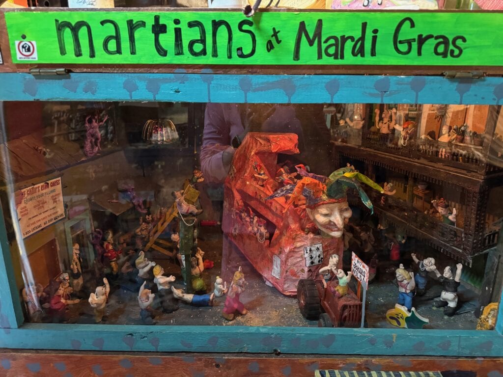 A colorful diorama titled Martians at Mardi Gras displays small figurines in festive poses, a large Mardi Gras float with a jester mask, and miniature buildings, all enclosed in a glass case with reflections.