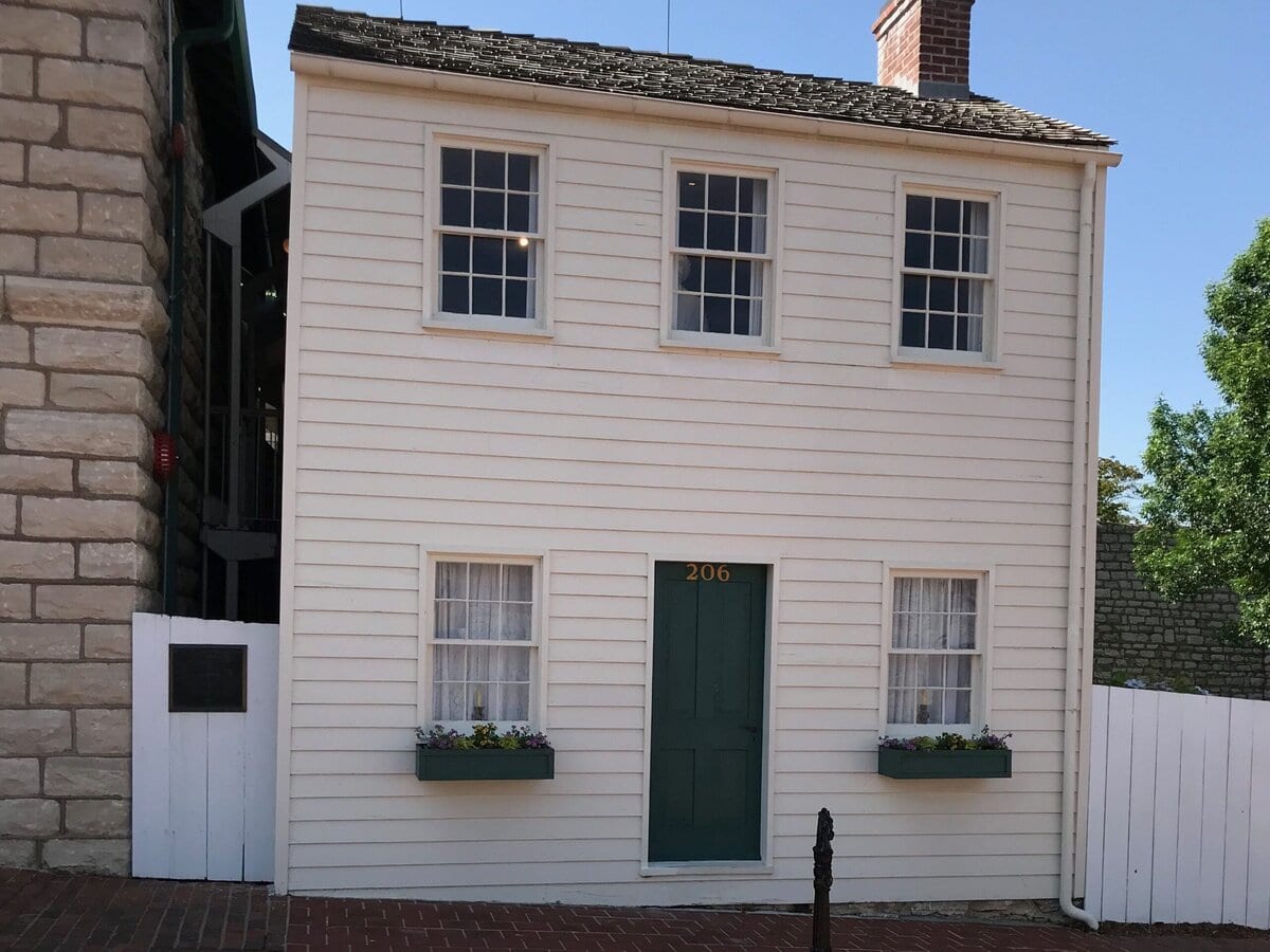 A small white two-story house with green door, flower boxes under the windows, and a brick sidewalk and street in the foreground, set against a sunny blue sky.