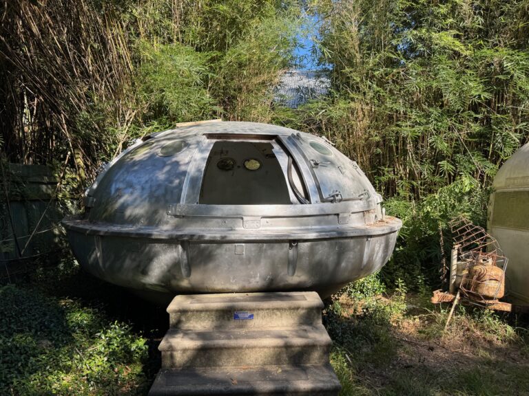 A gray, dome-shaped metal pod with a hatch and control dials sits on a concrete platform surrounded by dense green bamboo and plants in bright sunlight.