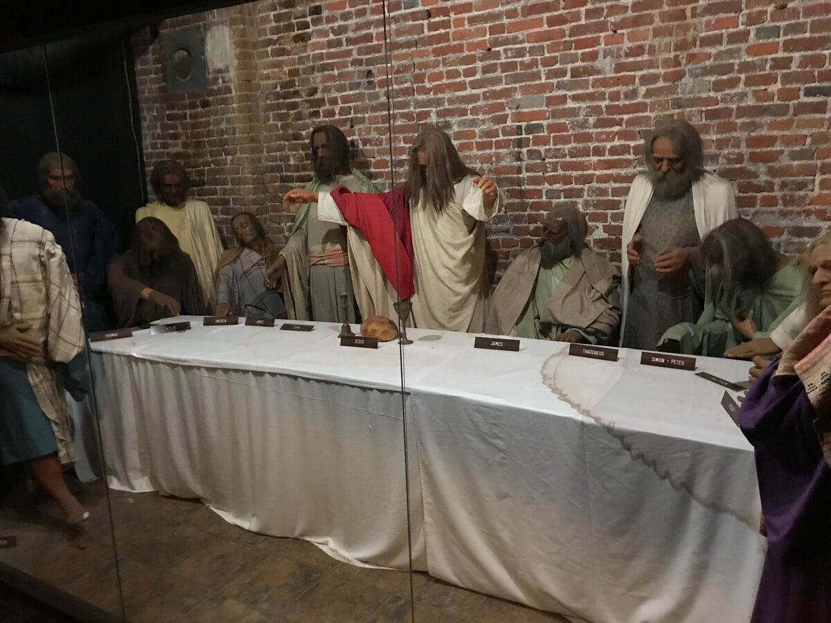 Wax figures depicting The Last Supper are arranged behind glass, with Jesus and his disciples gathered around a long table against a brick wall. Nameplates identify each figure.