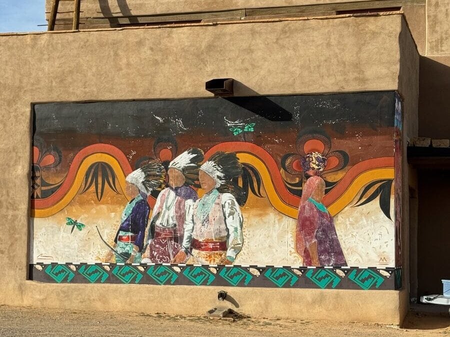 A mural on an adobe wall depicts four figures in traditional Indigenous attire with headdresses, colorful patterns, and two dragonflies, set against a vibrant background of orange, yellow, and black wavy lines.