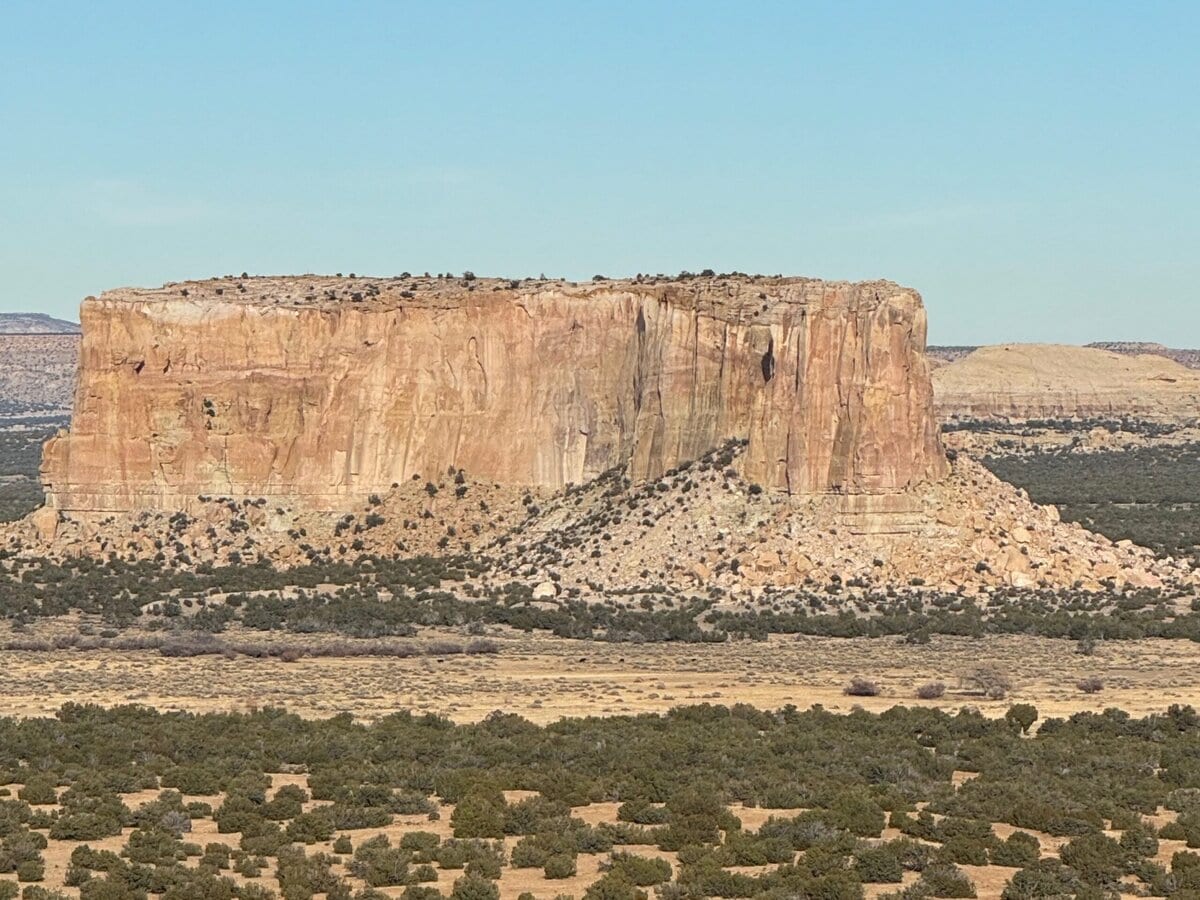 Large sandstone mesa rises above a flat, arid landscape dotted with shrubs and small trees under a clear blue sky.