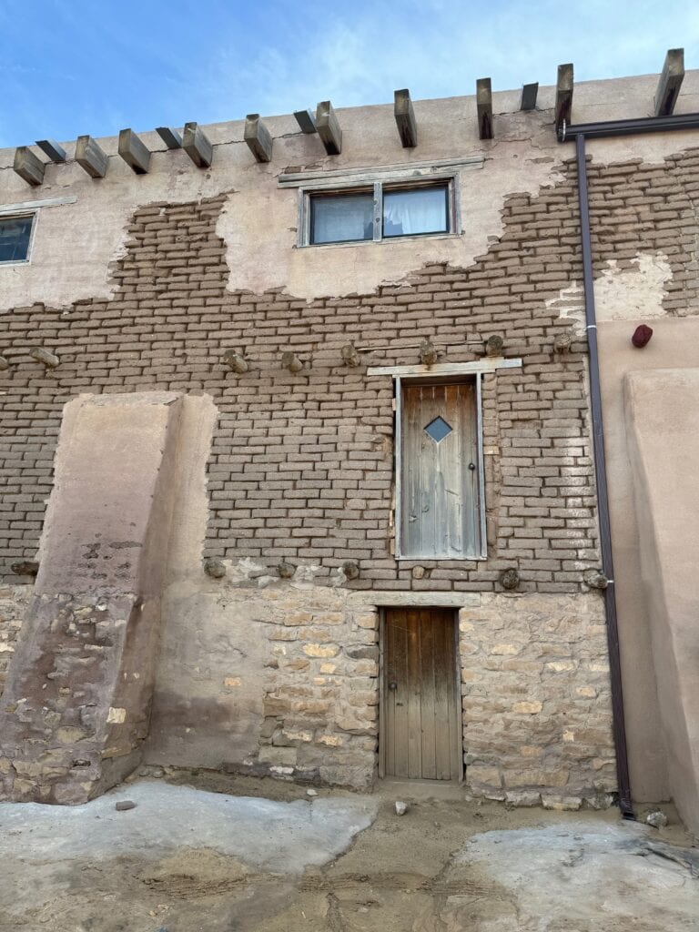An old, weathered building with adobe and brick walls features two doors, one on the ground level and one oddly placed above it with no stairs. A small window sits near the roof, and exposed wooden beams line the top.