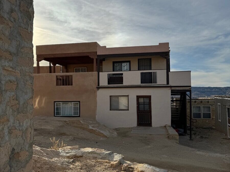 A two-story adobe-style building with balconies stands on a sandy, rocky landscape under a partly cloudy sky. A stone wall is visible in the foreground on the left side.