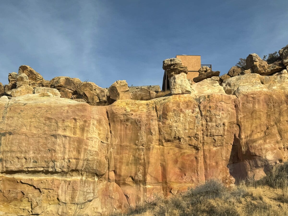 A tan, modern building sits atop a rugged sandstone cliff with streaks of yellow, orange, and red, under a clear blue sky. Sparse dry grass grows at the base of the rock formation.