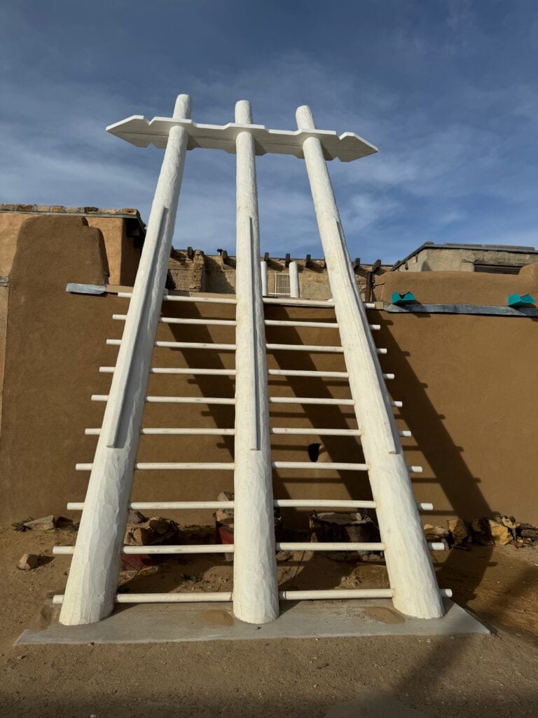A large traditional Pueblo ladder with three vertical poles and flat white steps leans against an adobe structure under a blue sky.