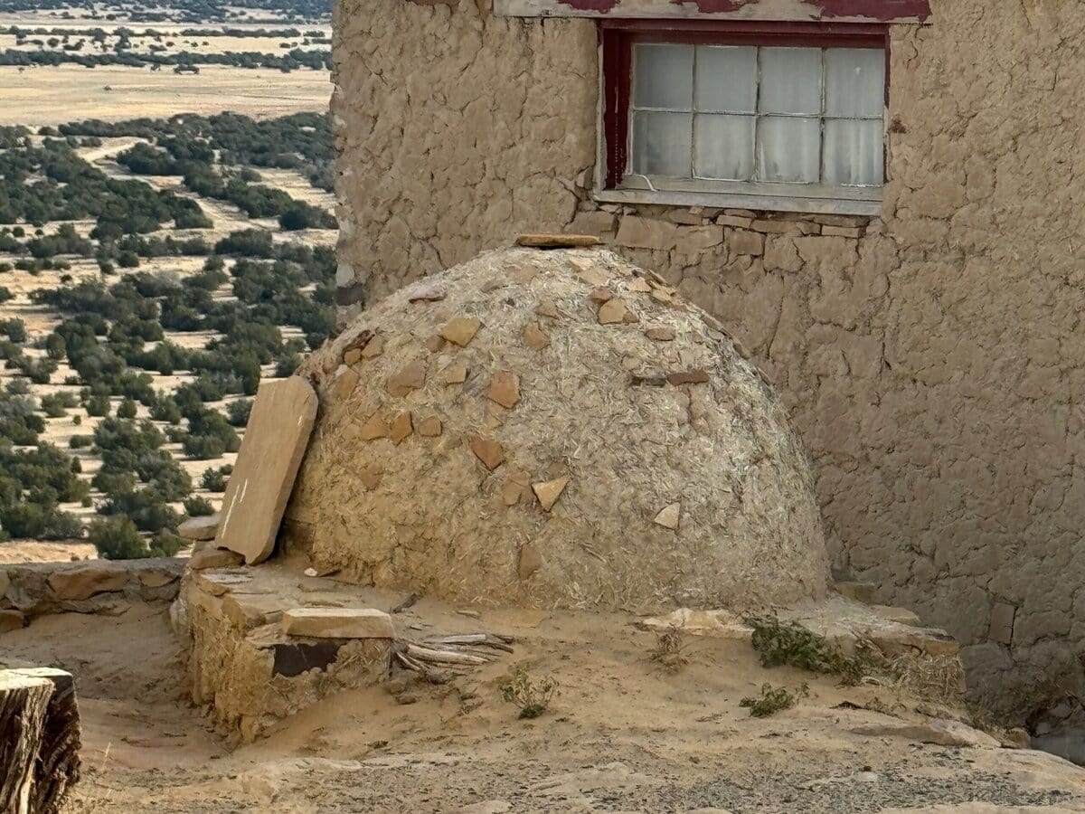 A dome-shaped adobe oven sits next to a weathered building with a red-framed window, overlooking a desert landscape with scattered trees and distant mesas.