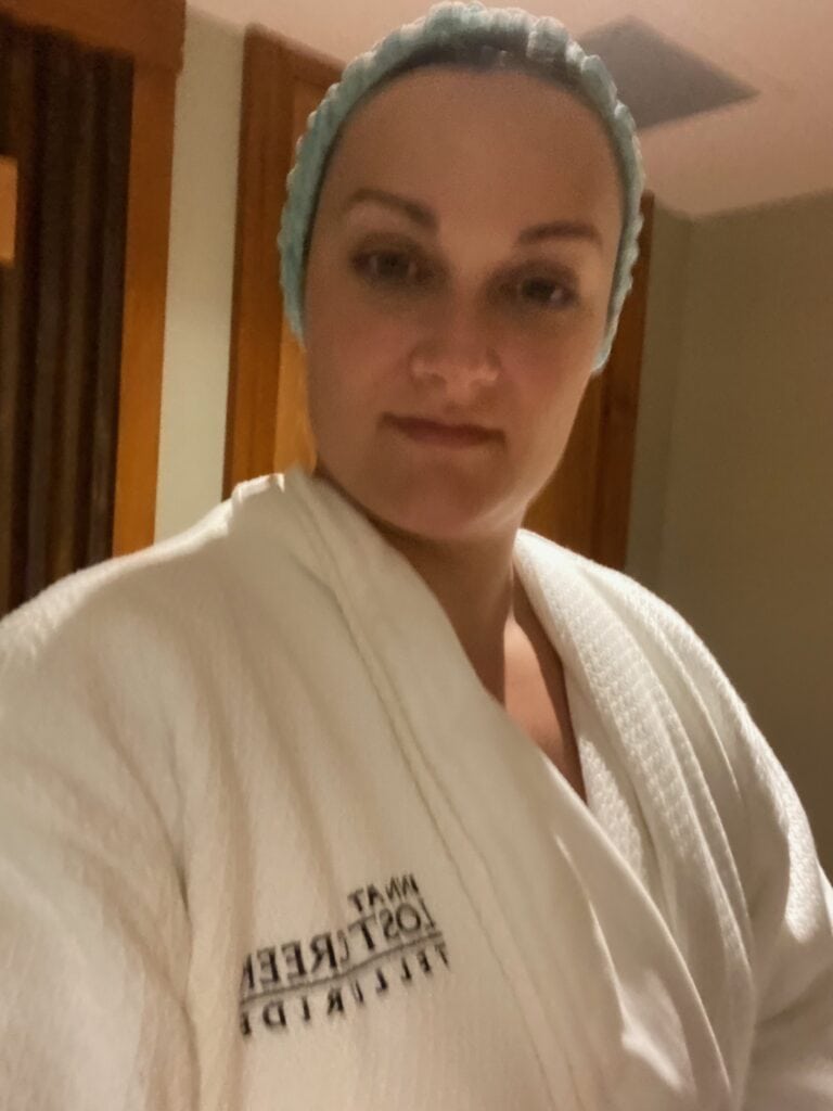 A person wearing a white spa robe and a blue hair cap stands indoors, looking at the camera. The robe has text on the left side. The background includes wooden doors and neutral-colored walls.