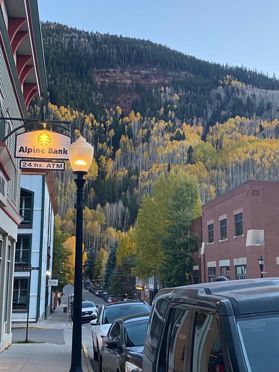 A street lined with buildings and parked cars leads toward a forested hillside covered in autumn trees with yellow leaves. A sign for Alpine Bank and a streetlamp are visible in the foreground.