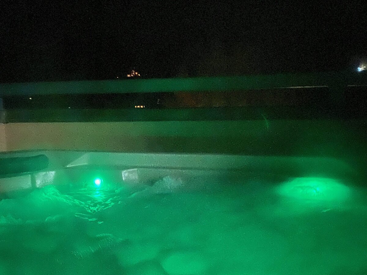 A bubbling hot tub glows with green underwater lights at night, surrounded by a dark background and a railing. Distant small lights are visible beyond the tub.