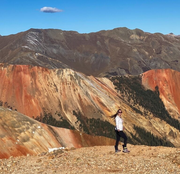 A woman wearing sunglasses and athletic clothing stands on rocky terrain with vibrant, multicolored mountains and a clear blue sky in the background. A single small cloud floats above the mountains.