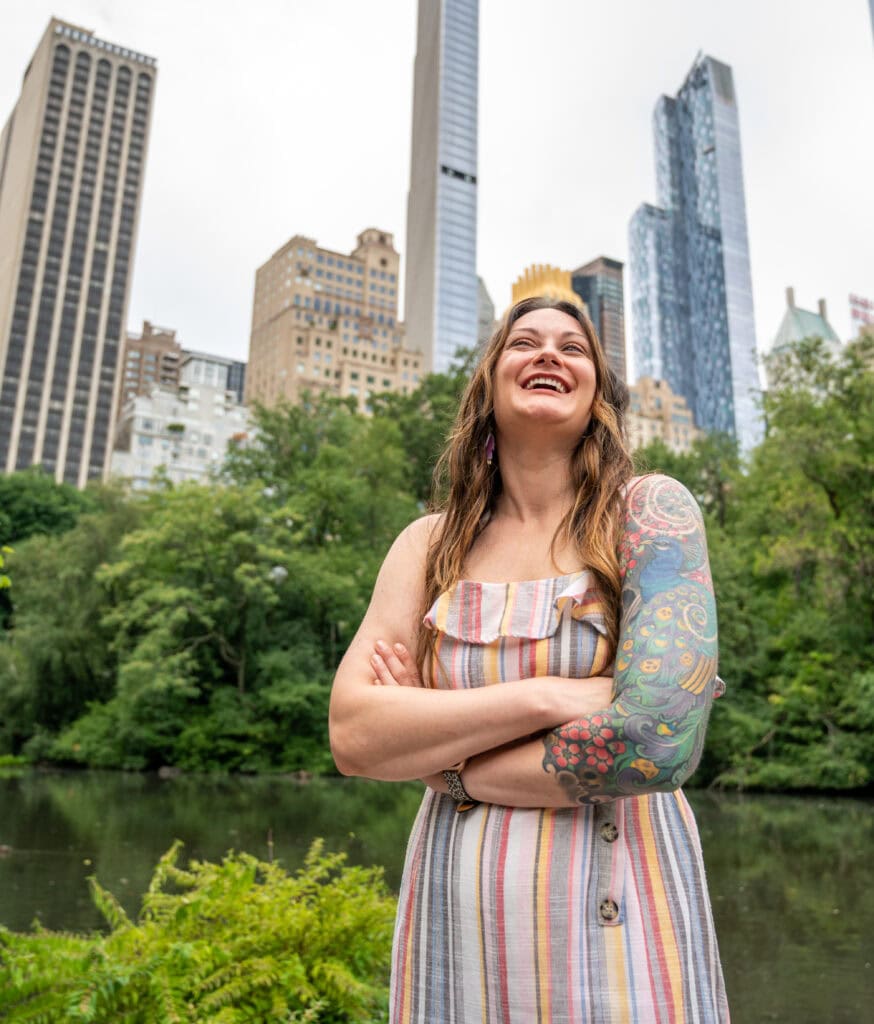 A smiling woman with long hair and a colorful sleeve tattoo stands with arms crossed in front of a pond and tall city buildings, surrounded by greenery.