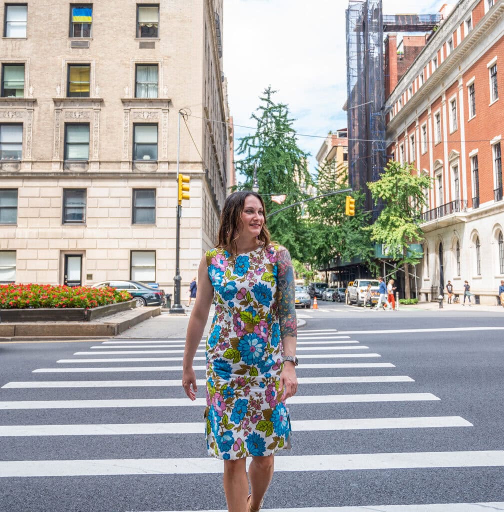 A woman in a colorful floral dress crosses a city street at a crosswalk, smiling. Tall buildings, trees, cars, and pedestrians are visible in the background on a sunny day.