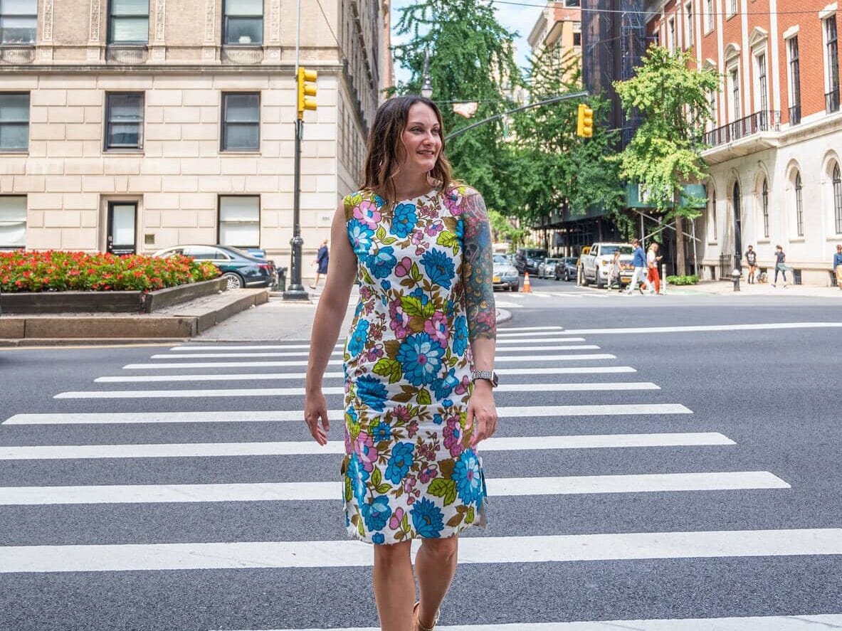 A woman in a colorful floral dress crosses a city street at a crosswalk, smiling. Tall buildings, trees, cars, and pedestrians are visible in the background on a sunny day.