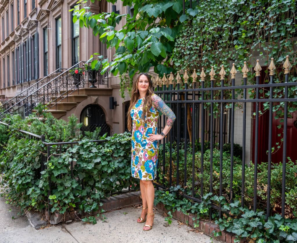 A woman in a colorful floral dress stands on a sidewalk next to a wrought iron fence covered in green ivy, with brownstone buildings and leafy plants in the background.