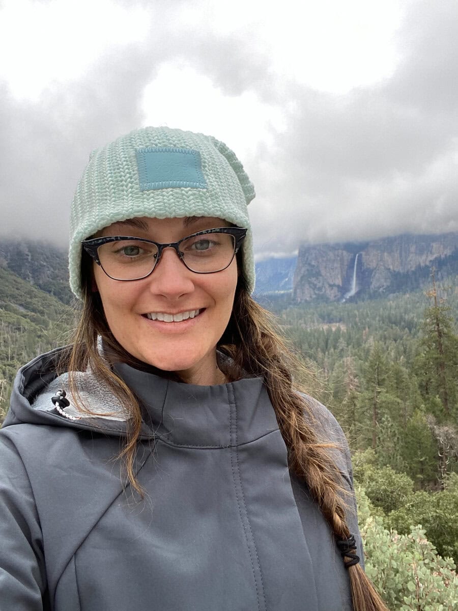 A woman wearing glasses, a gray jacket, and a knitted beanie smiles at the camera in front of a forested valley—one of the best spring destinations in the U.S.—with cloudy skies and a waterfall in the distance.