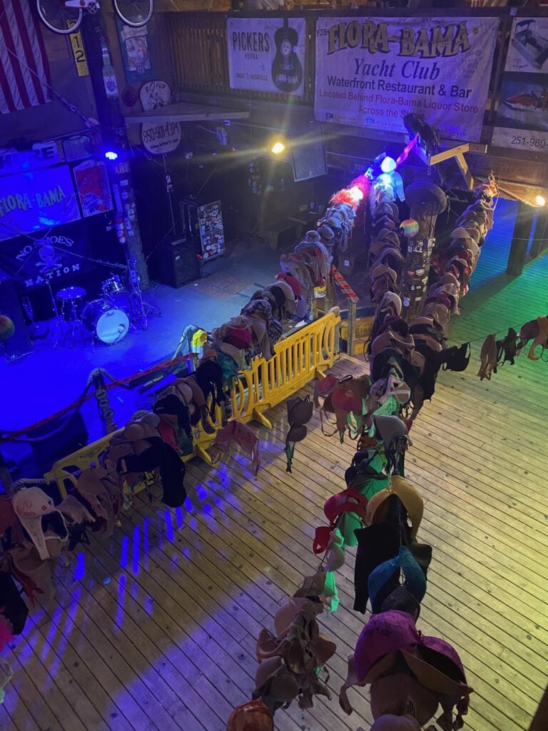 A wooden stage and dance floor at the Flora-Bama Yacht Club, with a drum set on stage and dozens of bras hanging from ropes and a yellow barricade, under colorful lights. Signs and banners decorate the background.