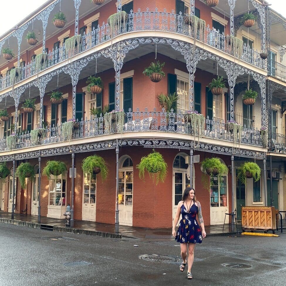 A woman in a blue dress walks across a wet street in front of a historic brick building with ornate ironwork balconies and hanging plants in New Orleans.