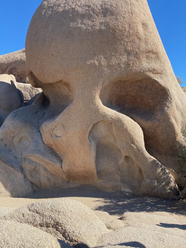 A large rock formation resembling a human skull, with deep eye sockets and a nose cavity, sits under a clear blue sky in a desert landscape.