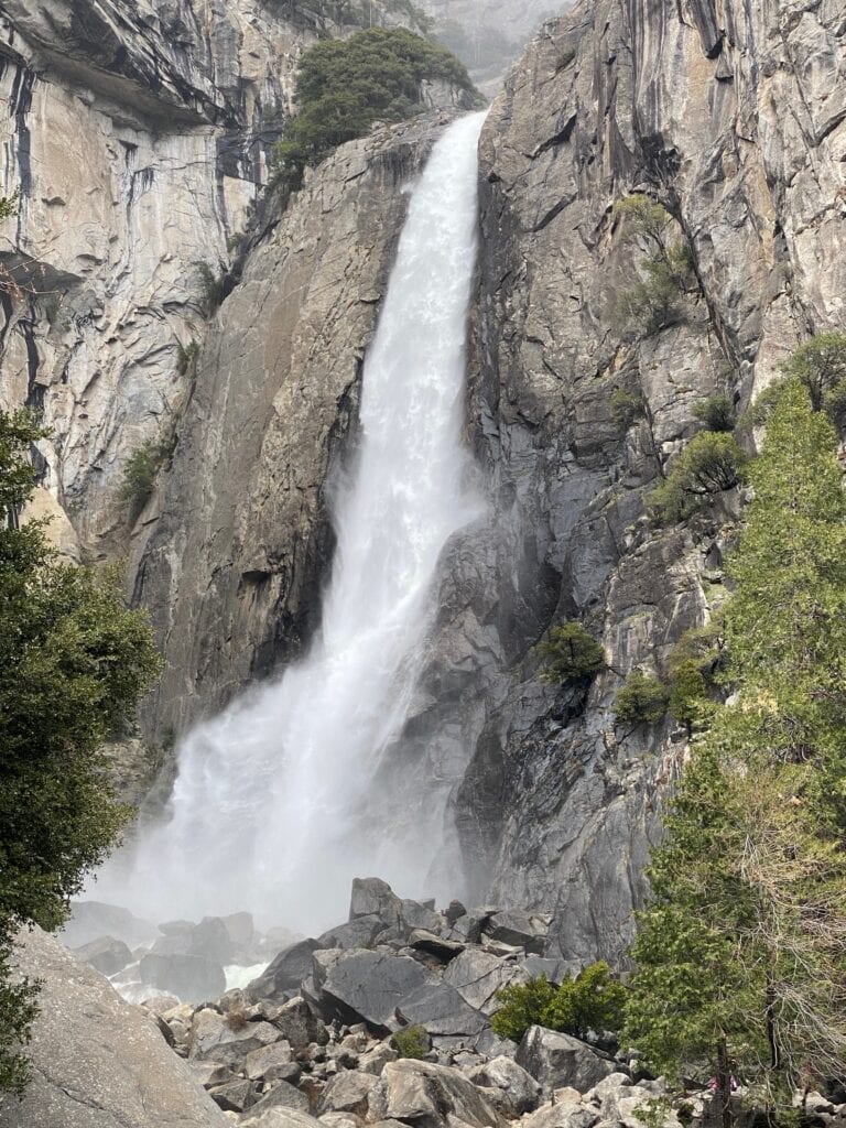 A tall waterfall cascades down a steep, rocky cliff surrounded by lush green trees and mist at the base. Large boulders are scattered around the waterfall’s plunge pool.