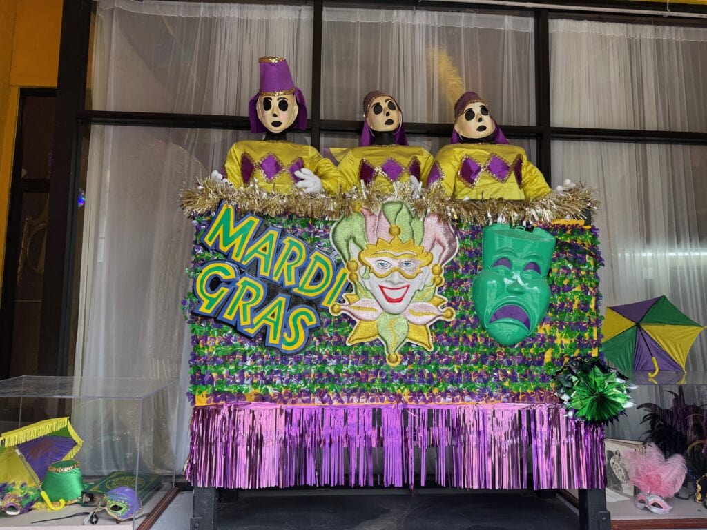A colorful Mardi Gras float with three mannequin figures in yellow, purple, and green costumes. The float is decorated with shiny fringe, masks, and a large “MARDI GRAS” sign in bold letters.