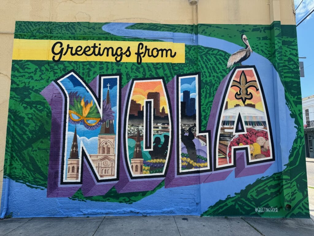 A vibrant mural reads Greetings from NOLA with each letter filled with New Orleans landmarks, cultural symbols, and a pelican above, set against a green and blue background.