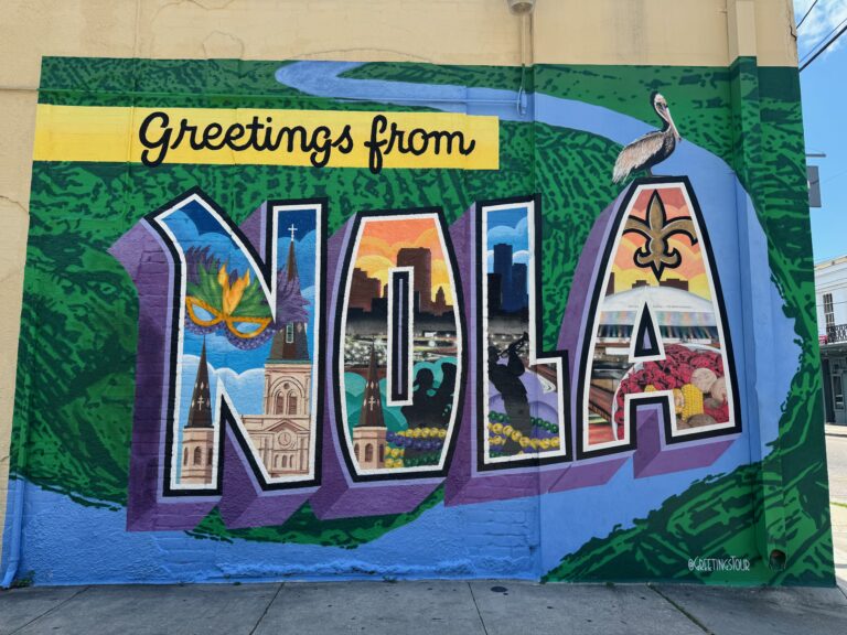 A vibrant mural reads Greetings from NOLA with each letter filled with New Orleans landmarks, cultural symbols, and a pelican above, set against a green and blue background.