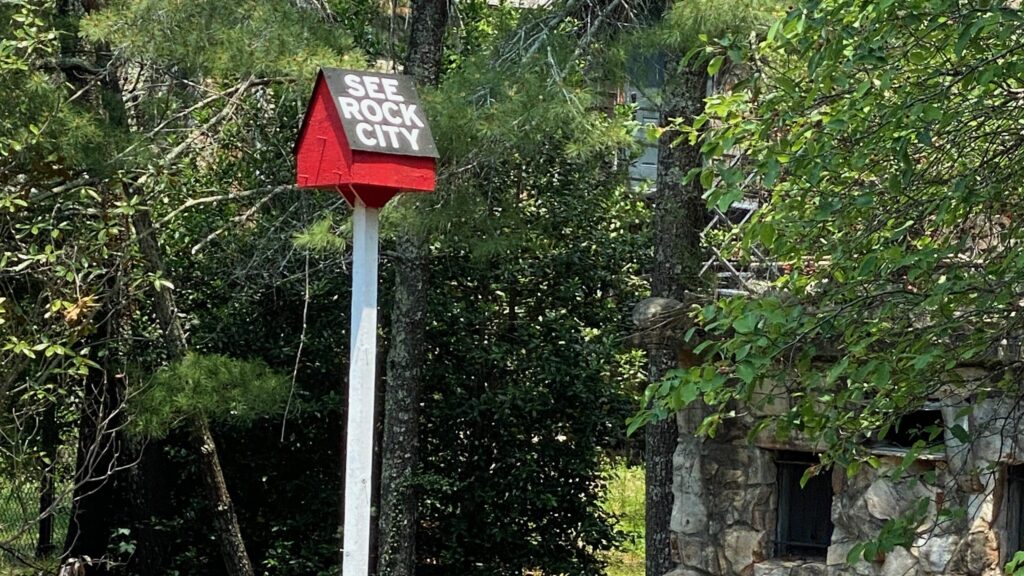 A small birdhouse painted red and black with the words See Rock City sits atop a tall white pole in a yard with stone landscaping, surrounded by trees and a rustic stone building in the background.