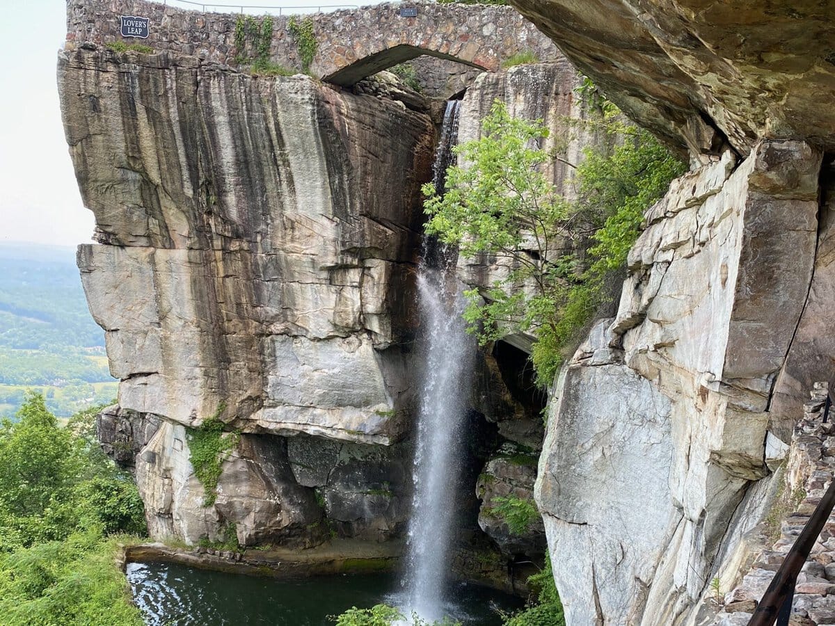 A waterfall cascades from a stone bridge into a pool below, surrounded by lush greenery and rocky cliffs, with a metal railing along a stone pathway in the foreground.