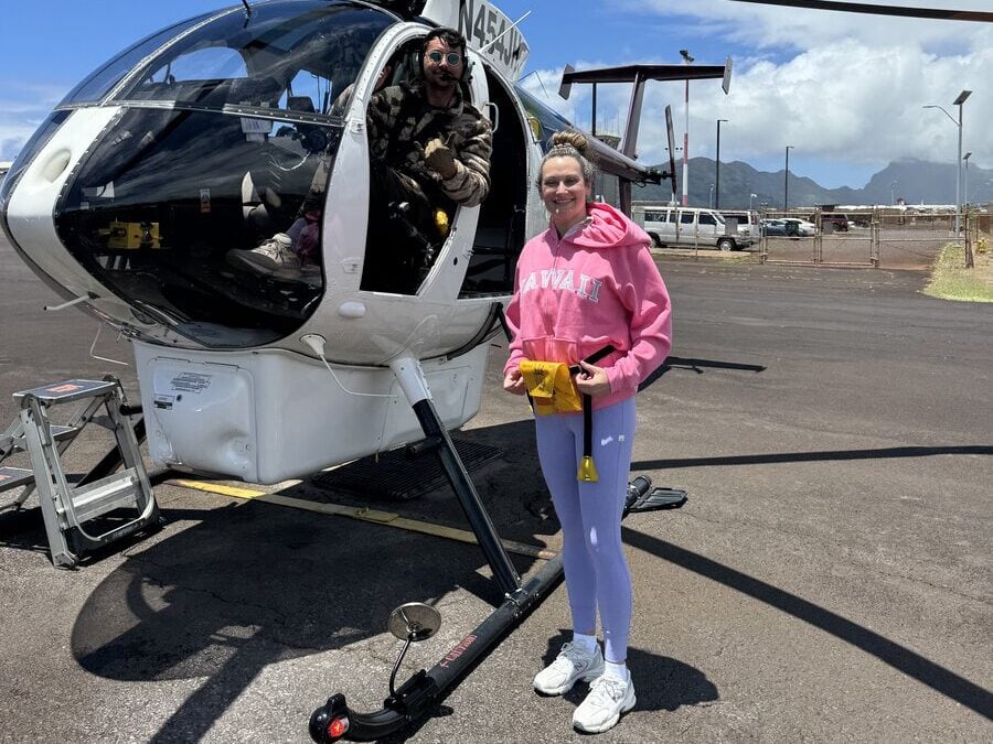 A woman in a pink Hawaii hoodie stands by a helicopter, holding a yellow item. A man sits inside the helicopter, smiling. The scene is outdoors with mountains and blue sky in the background.