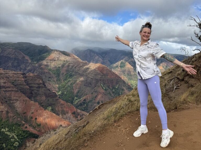 Person in purple leggings and white shirt stands smiling with arms outstretched on a mountain trail, with colorful, lush canyon hills and a partly cloudy sky in the background.