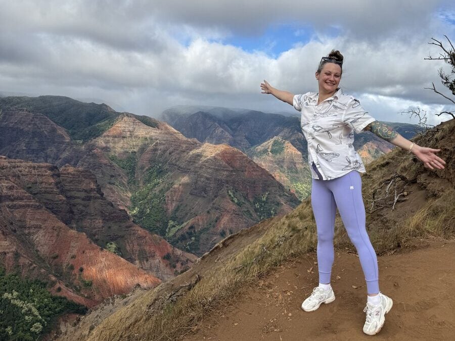 Person in purple leggings and white shirt stands smiling with arms outstretched on a mountain trail, with colorful, lush canyon hills and a partly cloudy sky in the background.