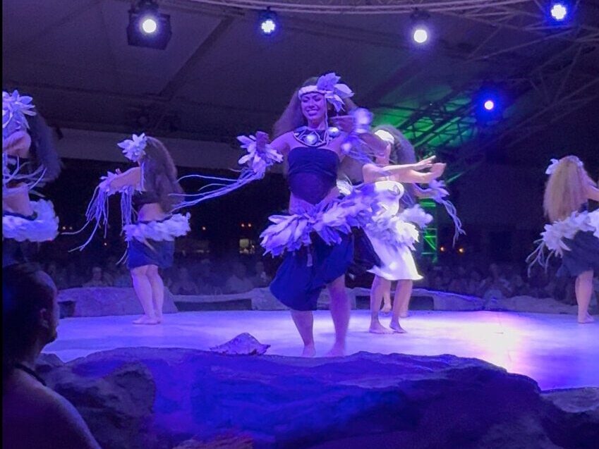 A group of dancers in traditional Polynesian attire perform on a stage under purple lighting, with an audience watching in the background. The main dancer wears a feathered headpiece and a grass skirt, bringing to life one of the unforgettable things to do on Kauai—experiencing an authentic island dance performance.
