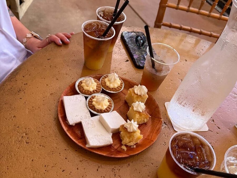 A round table with iced drinks in plastic cups, a water carafe, and a wooden plate holding small cakes, white rectangular slices, and tarts with whipped cream—perfect for relaxing after exploring things to do on Kauai. A phone and chair are visible in the background.